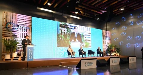 Conference stage with large screens displaying speakers, panel seating, and audience podium at corporate event
