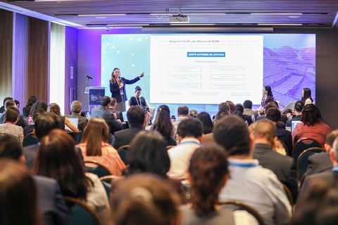 Speaker presenting to a large audience in a conference hall with purple lighting and projection screens displaying content
