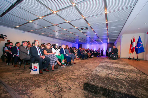 An audience seated in a modern conference hall with blue lighting and decorative flags visible on the right side of the room